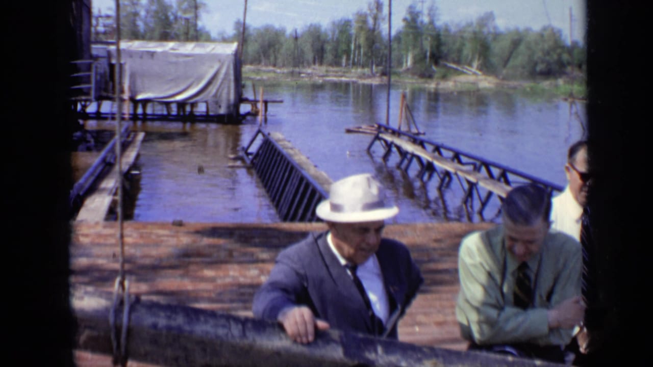 1971 Moscow home-movie frame of suited men at a lakeside railing, with a makeshift pier extending over the water.
