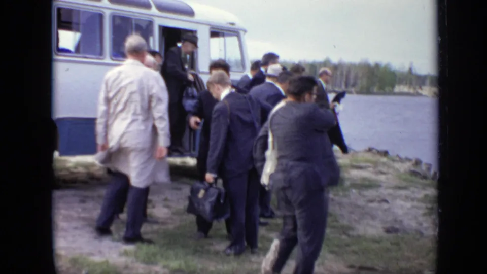 1971 Moscow 8mm frame showing suited men and briefcases approaching a white coach bus on a grassy lakeside shore.