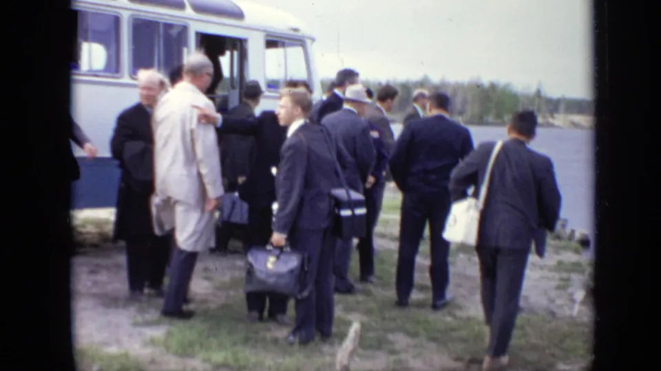 1971 Moscow home-movie frame of suited men carrying bags as they gather around an open white coach bus by the water.