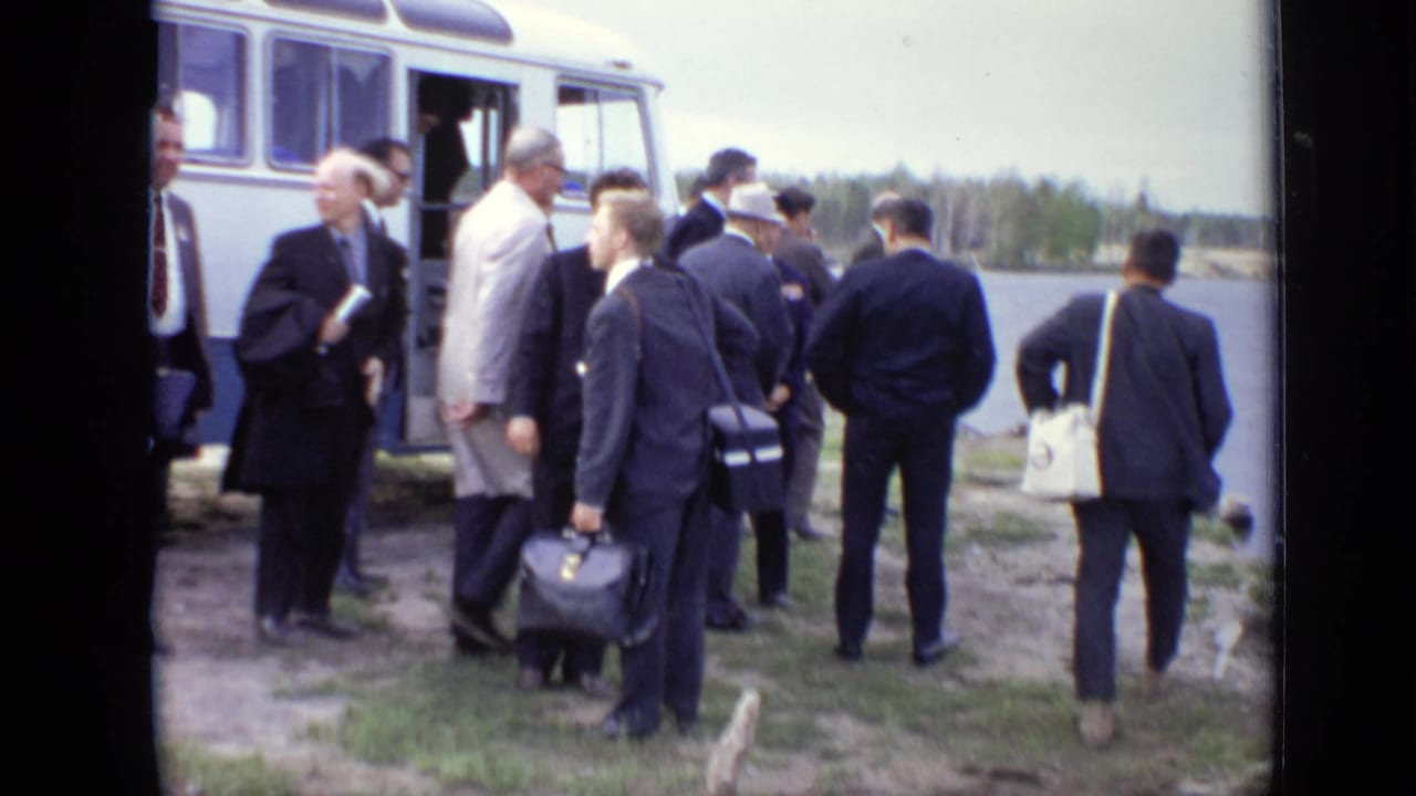 Men in suits stand beside a yellow-and-white vehicle near a lakeside.