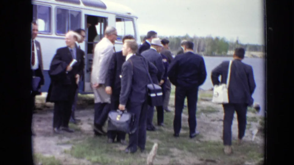 In Moscow-area 1971 home-movie footage, suited men with bags gather beside a yellow-and-white amphibious bus at the shore.