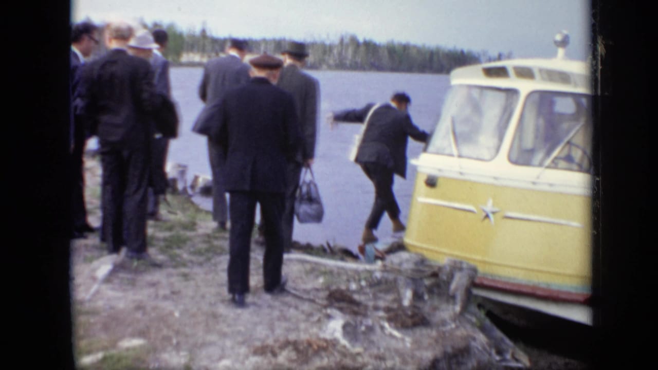 Men walk along a rocky shore beside the front of a yellow vehicle on the water.