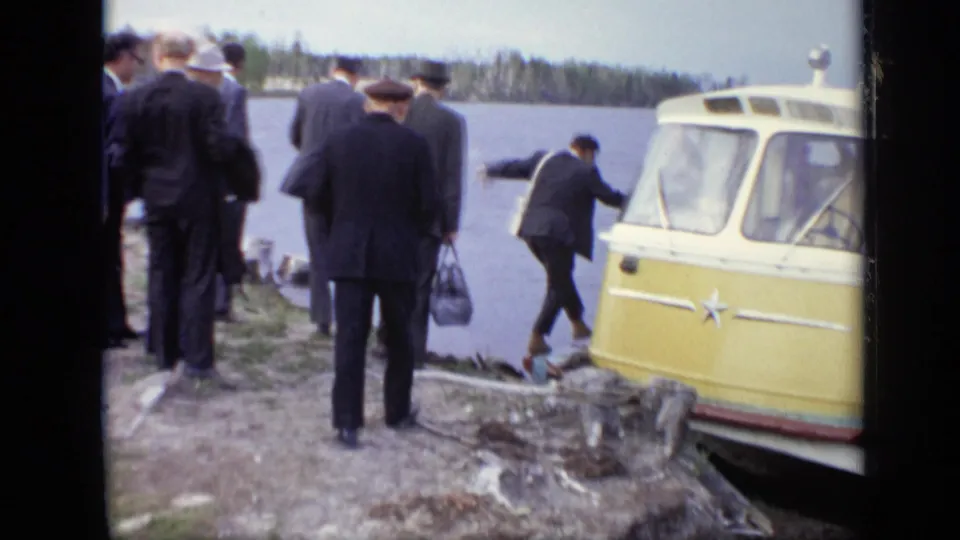 Men in formal clothing move along a rocky Moscow lakeshore beside the front of the yellow-and-white amphibious bus.