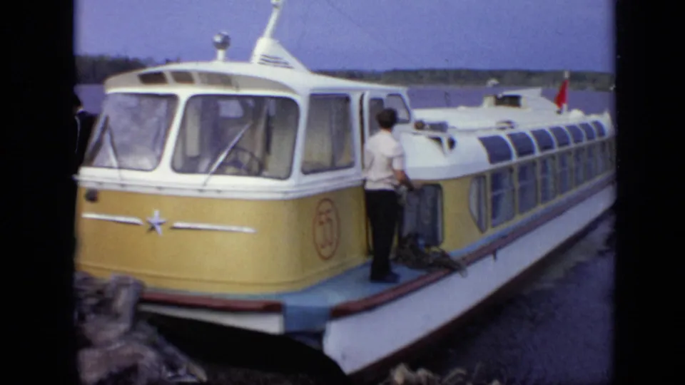A man stands beside the yellow-and-white amphibious bus or tour craft on calm water, with the lakeshore visible behind it.