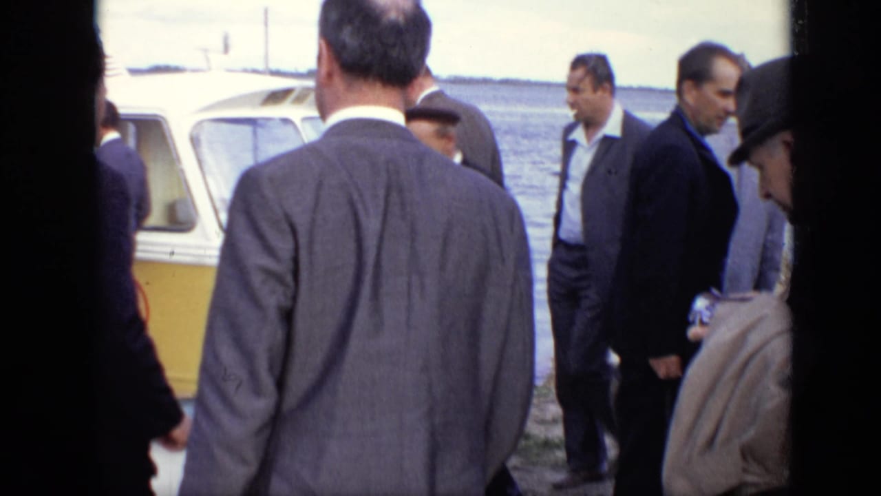 Men in suits stand near the docked yellow boat, with the water visible behind them.