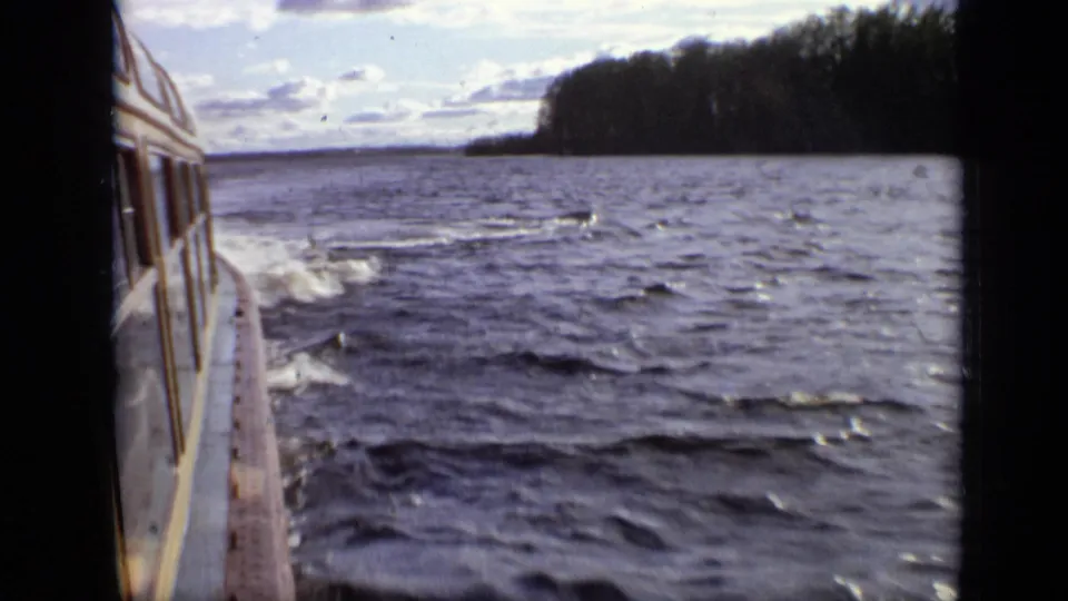 A handheld 8mm view from the boat shows choppy water, a low white wake, and a dark wooded shoreline beyond.