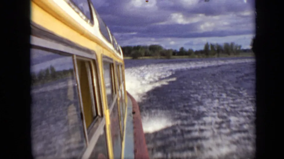The yellow-and-white cabin side is close to camera as the boat leaves a bright wake across the lake near a low shoreline.