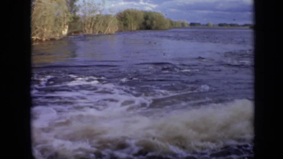The boat's wake churns across murky river water, with low wooded banks visible under a cloudy sky near Moscow in 1971.