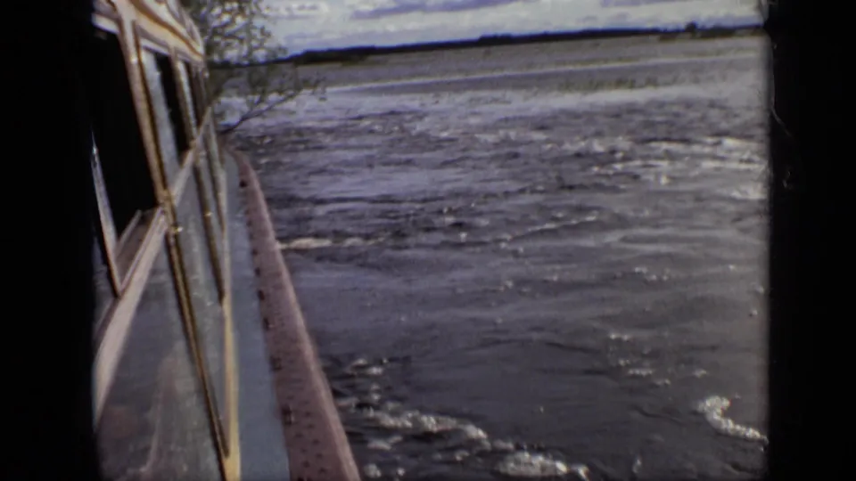 1971 home-movie view across rippling water with a shoreline in the distance and a windowed side edge at left.