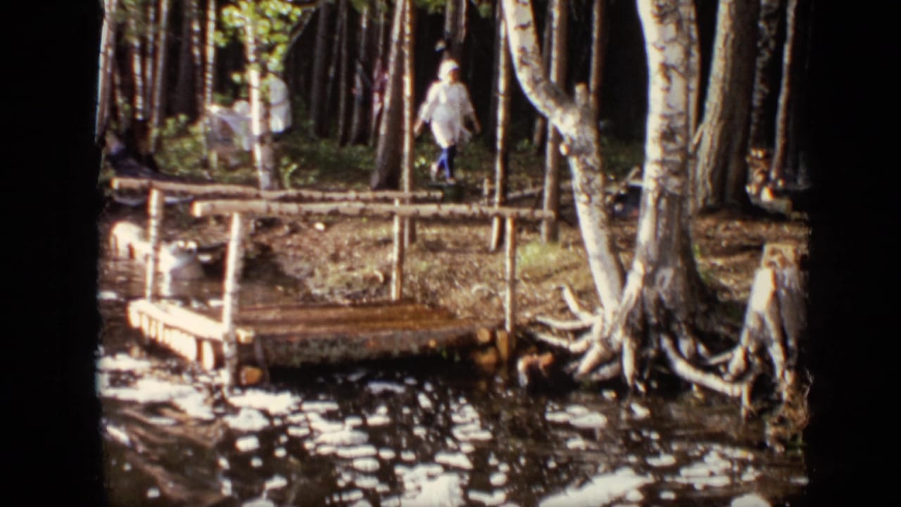 1971 Moscow home-movie frame with a woman walking quickly past trees near a handmade dock and shallow water.