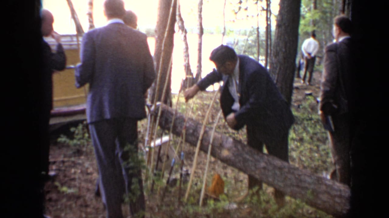 1971 Moscow 8mm frame showing suited men in a pine forest as one bends near sticks and a fallen log beside a bright lakeside background.
