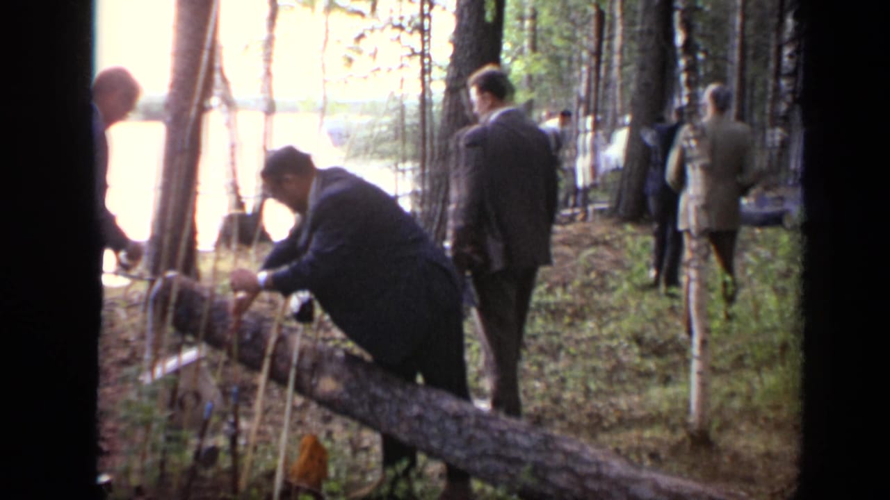 1971 Moscow 8mm frame with a suited man arranging kindling or sticks near a fallen log as other adults stand in the wooded area.
