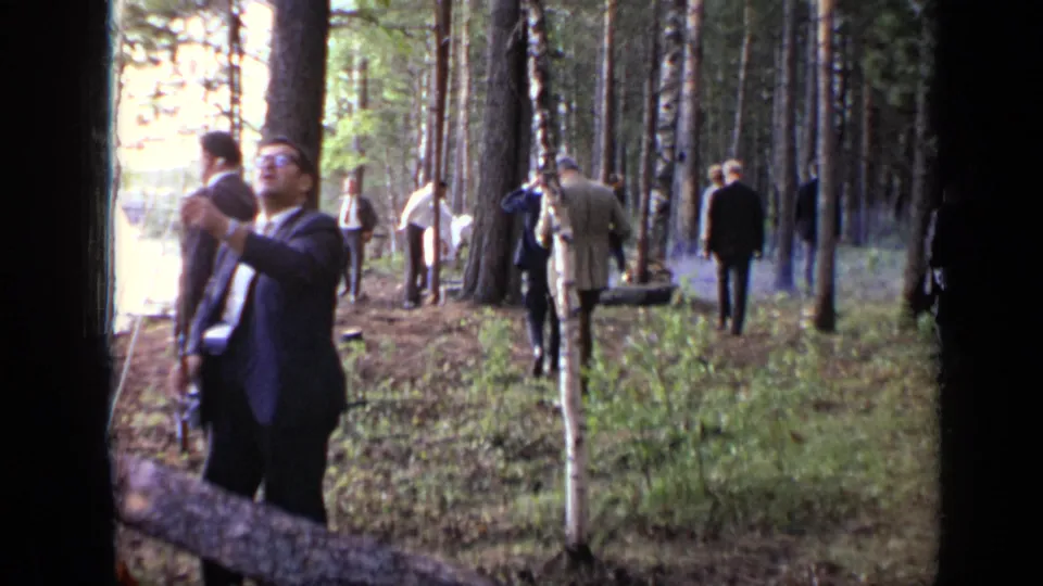 1971 Moscow 8mm frame showing suited adults in a pine forest, with one man near the foreground and smoke drifting from the wooded ground.
