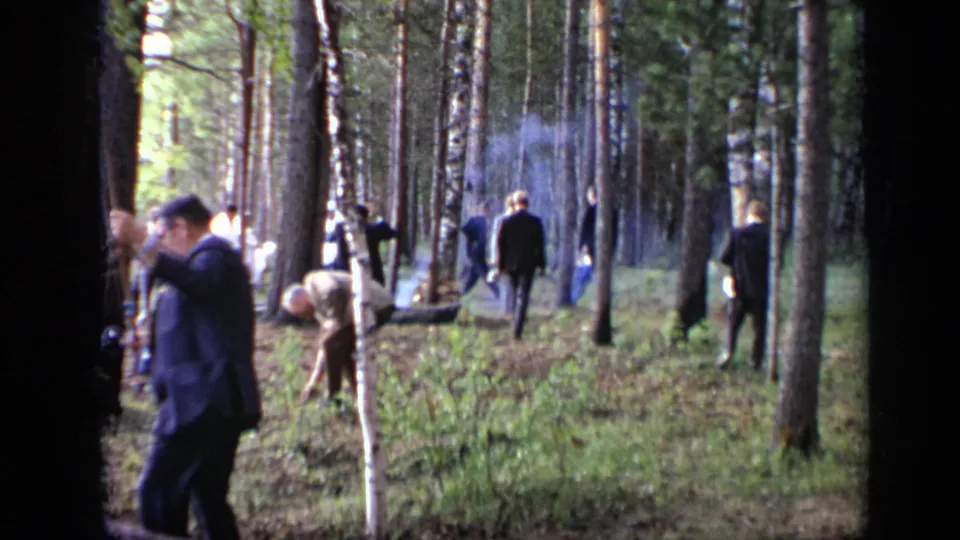 1971 Moscow 8mm frame of a suited man crossing a pine forest area with smoke, scattered adults, and a bright lakeside opening behind the trees.