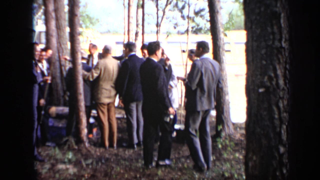 Men in suits standing among trees near a yellow-and-white boat.
