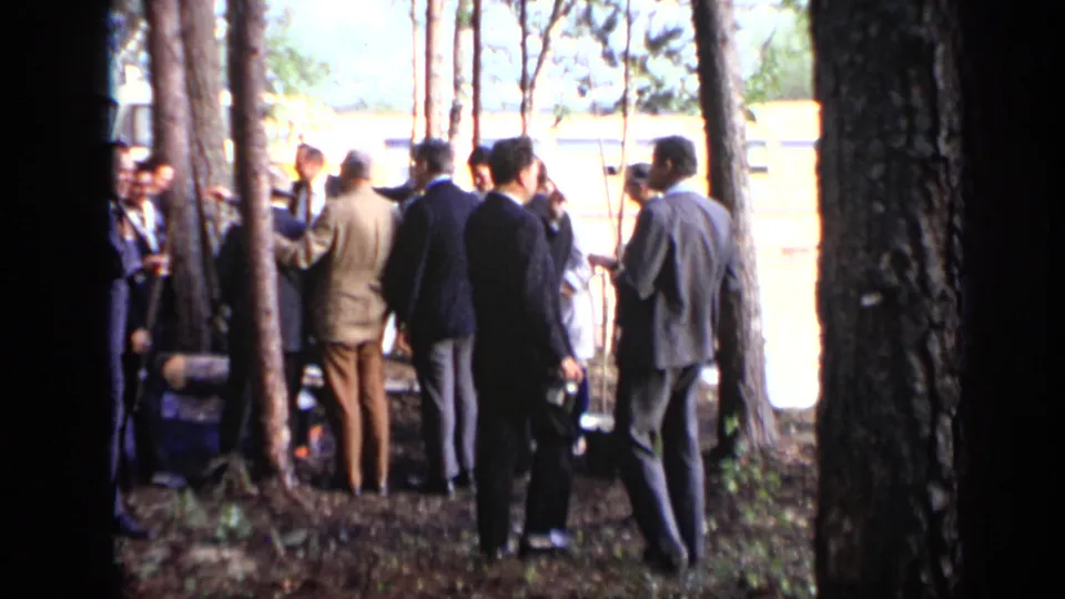 1971 Moscow home-movie frame of suited men gathered in a wooded area near a yellow-and-white riverboat.