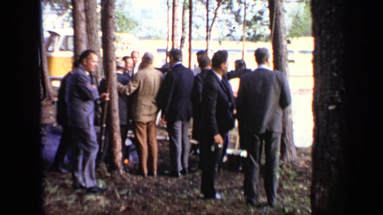 Suited men gather beside trees in Moscow, with a yellow-and-white riverboat visible behind them.
