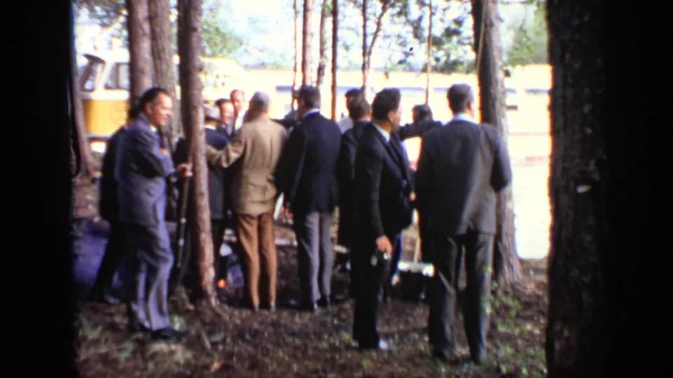 Suited men gather beside trees in Moscow, with a yellow-and-white riverboat visible behind them.