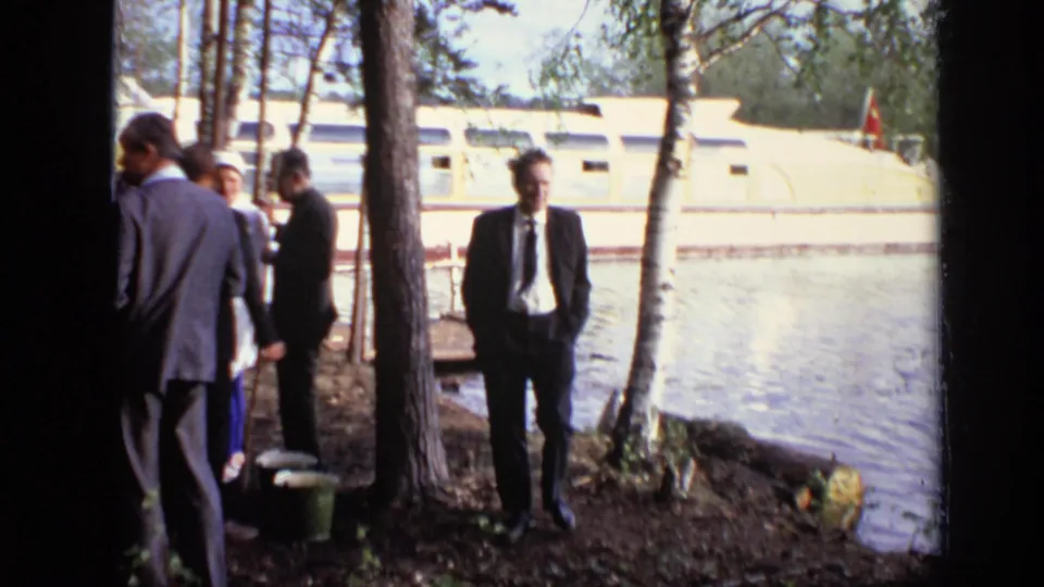 1971 Moscow frame showing a suited man by the water while other guests stand near trees and a riverboat.