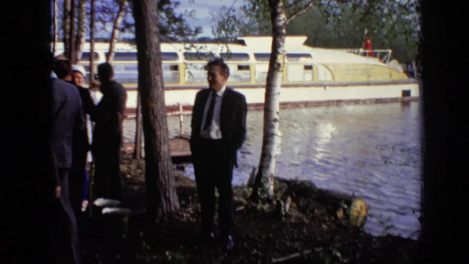 A suited man stands near trees along the water in Moscow, with a moored riverboat behind him.