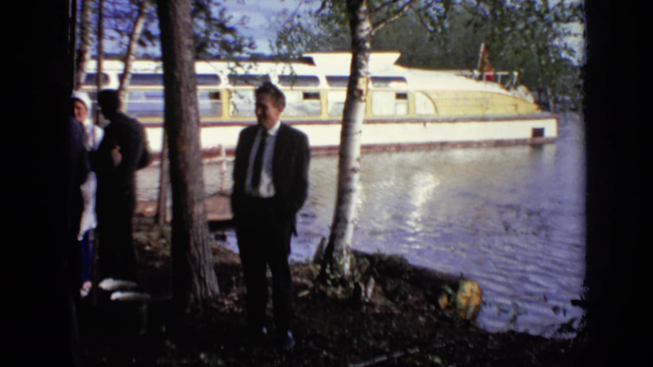 Men in suits stand on a wooded riverbank with a large houseboat moored behind them.