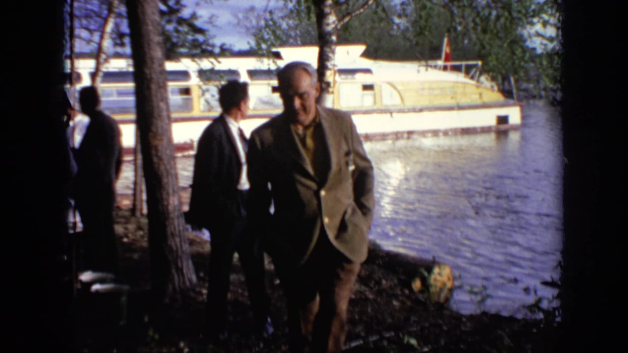 1971 Moscow home-movie frame with an older suited man approaching the camera as others stand by the water and houseboat.