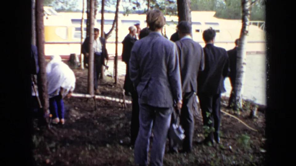 1971 Moscow riverside frame showing suited men observing a small campfire near trees, with the houseboat partly visible beyond.