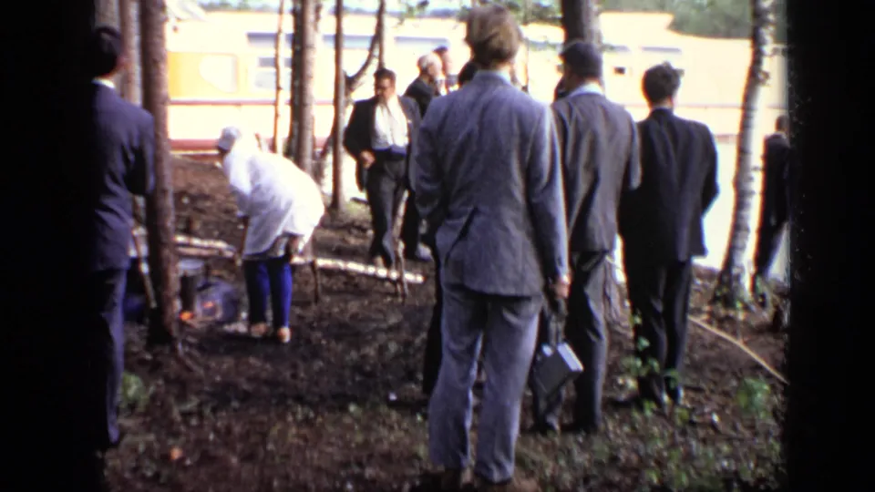 1971 Moscow home-movie frame of suited adults gathered around a small riverside fire, with the moored houseboat in the background.