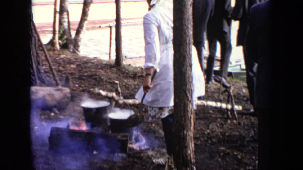 Smoking pots sit over a fire while adults stand among trees nearby.