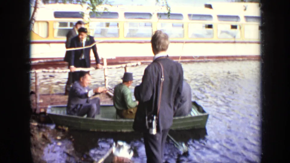 1971 Moscow home-movie frame showing suited adults around a small rowboat by water, with a yellow rail vehicle in the background.