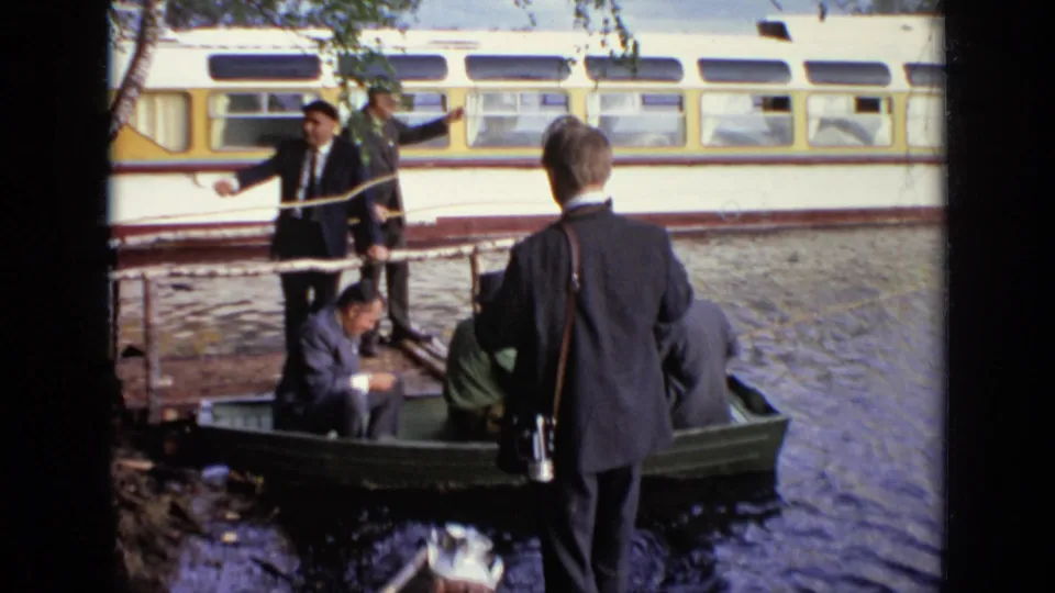 1971 Moscow home-movie frame with men in suits around a rowboat at the water's edge, a yellow rail vehicle visible behind them.