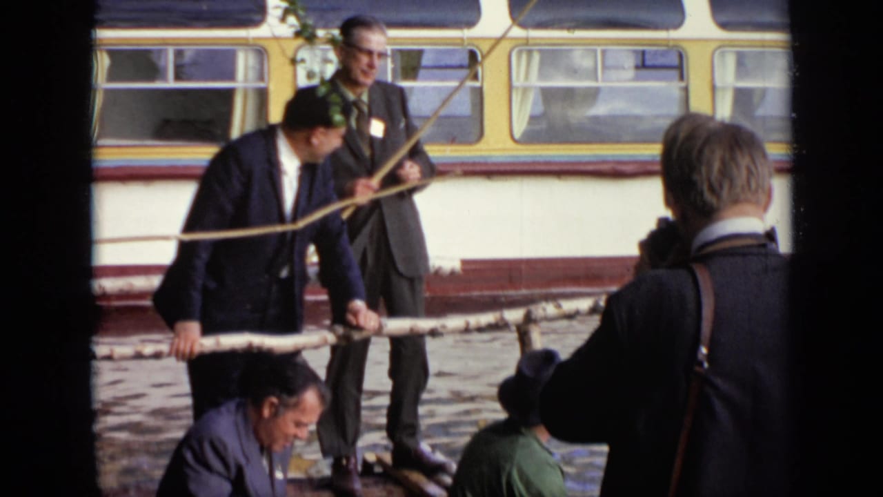 1971 Moscow lakeside frame with several adult men in suits, one holding a long branch or pole near a small boat and bus.