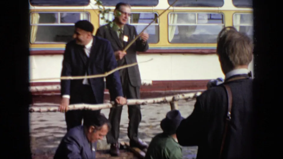 1971 Moscow home-movie frame showing a suited man leaning at the lakeshore while others sit nearby, with the yellow-and-white bus in the background.