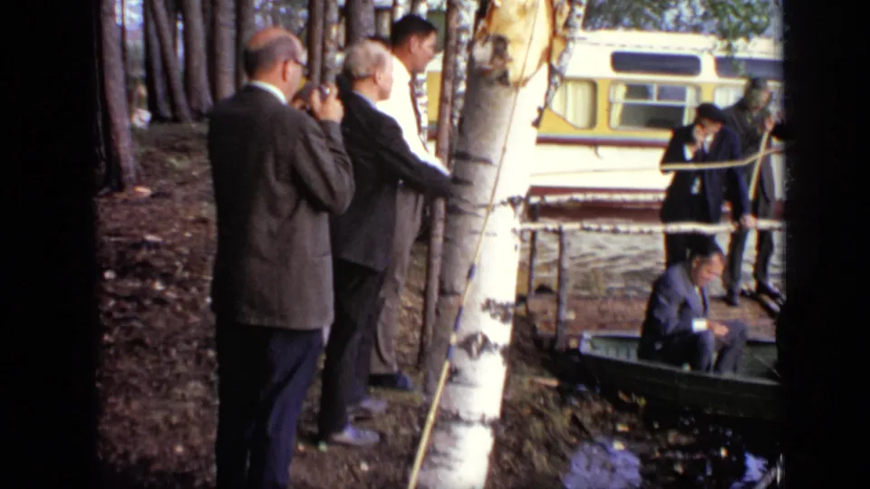 1971 Moscow 8mm frame with suited men watching from a wooded shore as others sit in a green boat beside a yellow-and-white bus.
