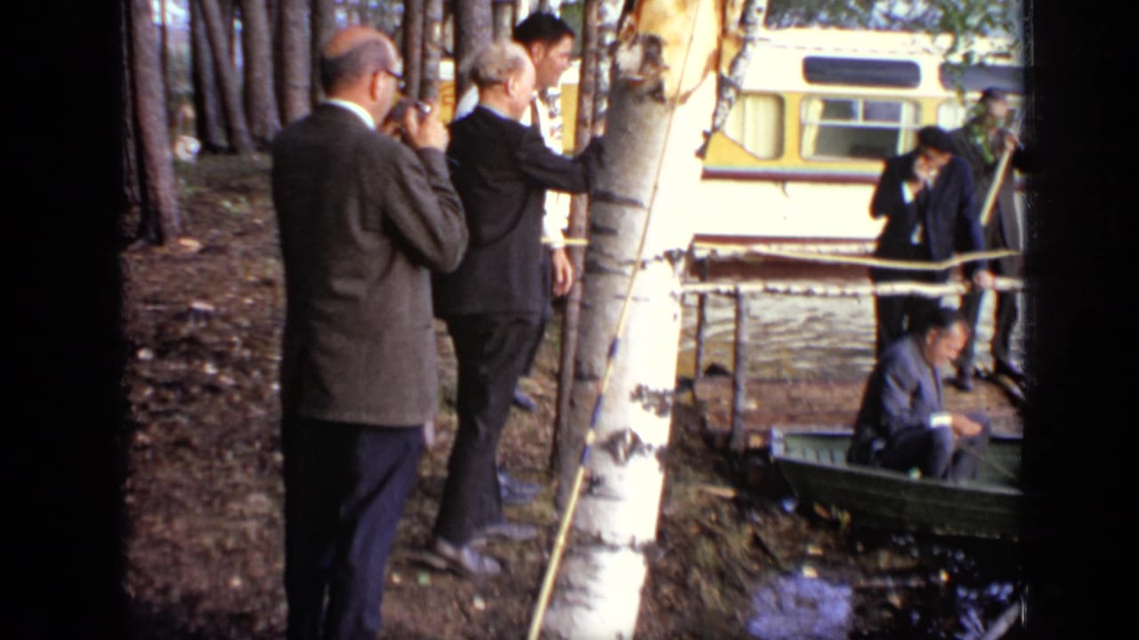 Men in suits stand by a birch tree near water, with a bus and small boat behind them.