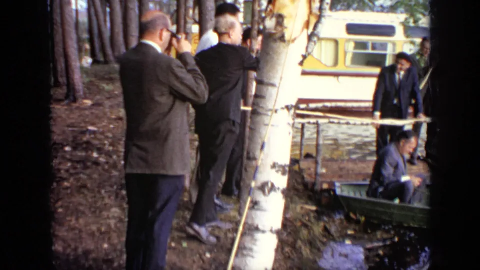 1971 Moscow 8mm frame with men in suits at a birch trunk, a man by a small boat, and a bus parked beyond the trees.