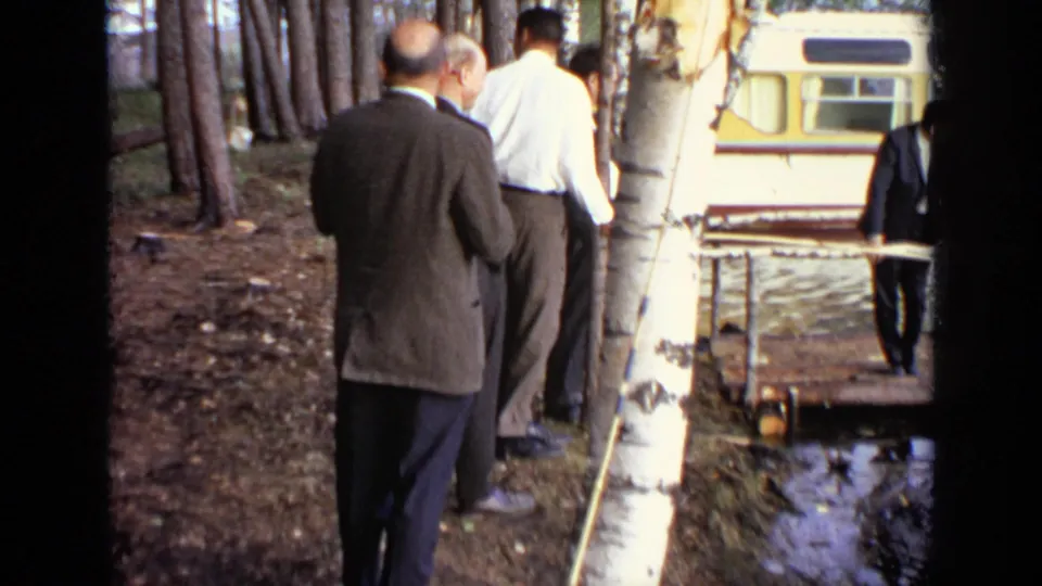 1971 Moscow 8mm frame showing suited men moving through a wooded bank beside a birch tree, small boat, and parked bus.
