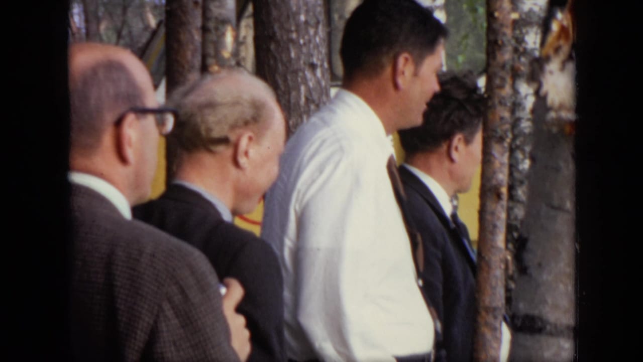 Older men in suits stand among birch trees near a yellow bus.