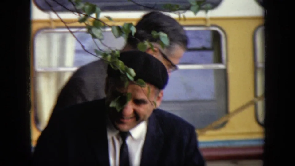 A suited man in a black beret moves past leafy branches with a yellow bus behind him in the Moscow 1971 footage.