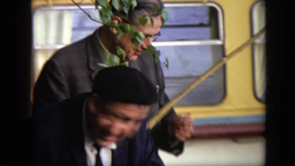 Two suited men stand close to a yellow bus, with one wearing a black beret and foliage crossing the foreground.