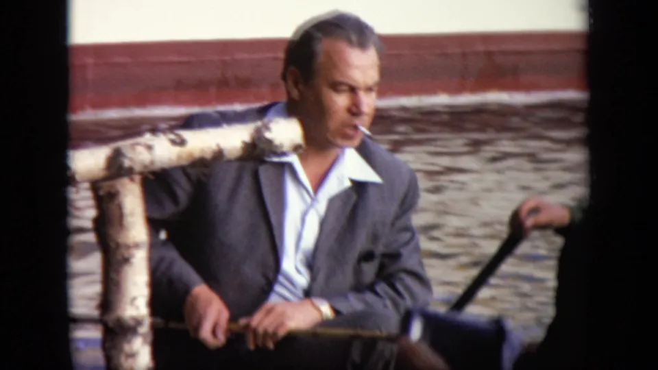 A suited man smokes while seated beside water and a rough wooden railing in the 1971 Moscow home-movie footage.