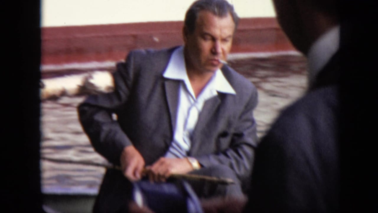 Man in a suit smoking in a rowboat, with another figure partly blocking the foreground.