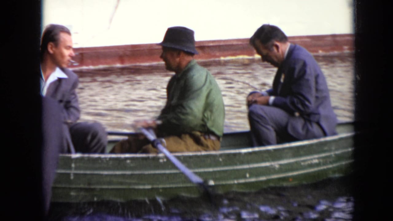 Three men sit in a green rowboat, with one man holding oars on the water.