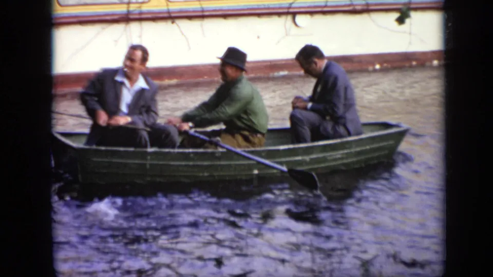 A small green rowboat carries three adult men past a yellow-and-white building wall in 1971 Moscow home-movie footage.