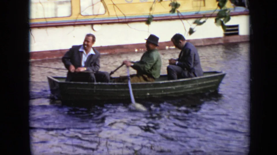 Three adult men sit in a green rowboat on a Moscow waterway, with an oar in motion and branches above the frame.