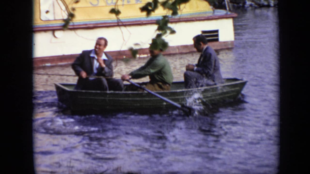 1971 Moscow home-movie frame showing three adult men in a green rowboat, with one rowing and a yellow-and-white riverside structure behind them.
