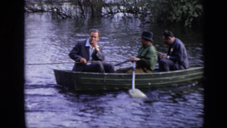 1971 Moscow river footage frame showing three adult men seated in a green rowboat, one holding an oar while the others sit quietly.