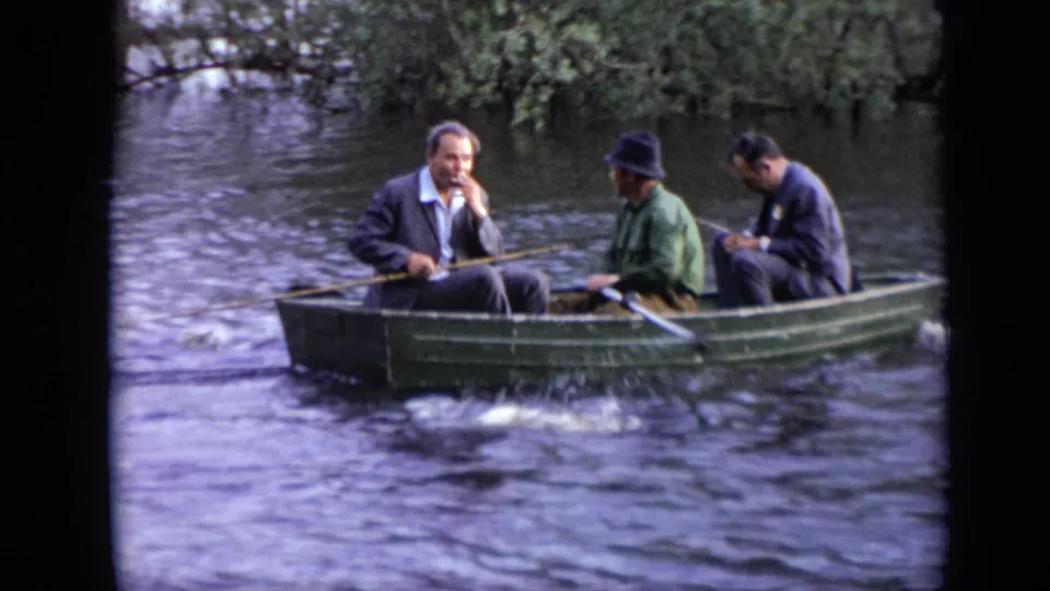 A 1971 Moscow home-movie frame shows three adult men in a green rowboat moving past a tree-lined riverbank.