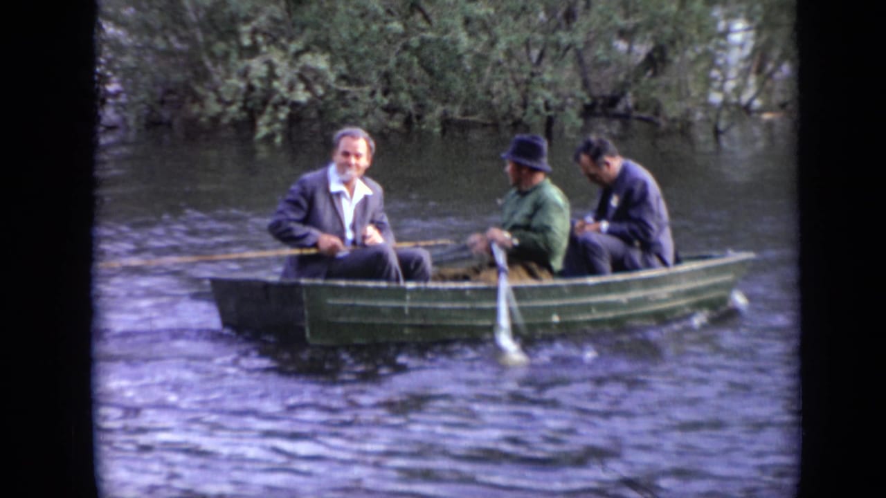 Three adult men sit in a green rowboat near trees on a river.