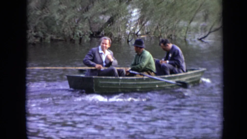 Three men in a green rowboat on a Moscow waterway, one rowing and another seated at the bow in a dark suit.